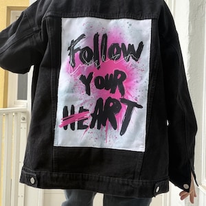 Könnte beinhalten: Schwarze Jeansjacke mit einem großen weißen Aufnäher auf dem Rücken. Der Aufnäher zeigt die Worte "Follow Your Heart" in schwarzer und rosa Schrift mit einem rosa Spritzdesign. Die Jacke hat ein klassisches Knopfdesign.