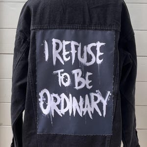 Puede incluir: Chaqueta vaquera negra con un parche rectangular en la espalda. El parche presenta el texto blanco "I REFUSE TO BE ORDINARY" en una fuente de estilo grafiti. La chaqueta se muestra en un maniquí.