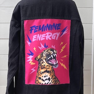 May include: Black denim jacket with a vibrant pink patch featuring a roaring leopard and the words "FEMININE ENERGY" in blue and pink. The jacket has a classic design with button closures and a collar.