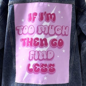 May include: A denim jacket with a light pink patch on the back. The patch features the text "IF I'M TOO MUCH THEN GO FIND LESS" in bubble letters, with a lightning bolt and star accents. The jacket is a medium wash.
