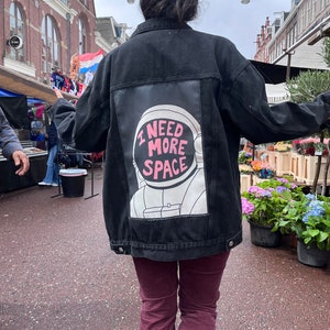 Può includere: Una persona indossa una giacca di jeans nera con una toppa di astronauta bianca sul retro. La toppa ha il testo "I NEED MORE SPACE" in lettere rosa.