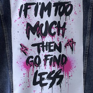 Puede incluir: Una chaqueta vaquera con un parche blanco cosido en la espalda. El parche tiene salpicaduras de pintura rosa y texto negro que dice "Si soy demasiado, entonces ve a buscar menos".