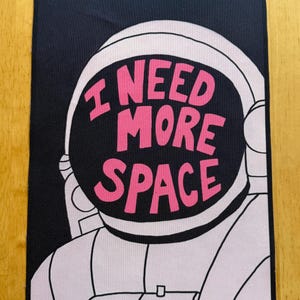 Könnte beinhalten: Schwarzer rechteckiger Aufnäher mit einem weißen Astronautenhelm und dem pinkfarbenen Text "I NEED MORE SPACE". Das Visier des Astronauten ist schwarz. Der Aufnäher ist wahrscheinlich aus Stoff und zum Aufnähen oder Aufbügeln auf Kleidung oder Taschen geeignet.