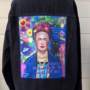 Puede incluir: Chaqueta vaquera negra con un retrato colorido de Frida Kahlo en la espalda. La obra de arte incluye flores, un gato negro y las palabras "Queen" e "Icon". La chaqueta se exhibe en un maniquí de madera.