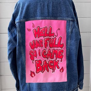May include: A blue denim jacket with a pink patch on the back. The patch features the phrase "HELL WAS FULL SO I CAME BACK" in red, stylized lettering. The jacket is displayed on a white background.