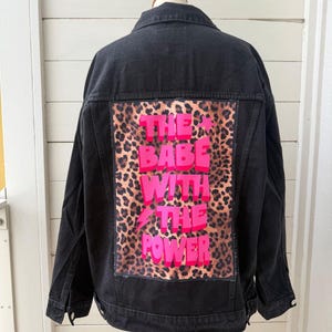 Op de afbeelding: Een zwart spijkerjack met een grote rechthoekige patch op de achterkant. De patch heeft een bruin en zwart luipaardprint patroon met de felroze tekst "THE BABE WITH THE POWER" en een kleine roze bliksemschicht.