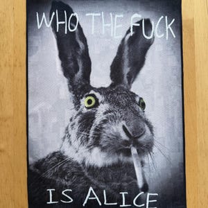 May include: A rectangular fabric patch featuring a black and white photograph of a hare with bright yellow-green eyes and a cigarette in its mouth. The text WHO THE FUCK IS ALICE is printed in white capital letters at the top and bottom edges.