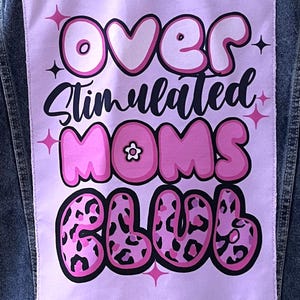 May include: A denim jacket with a light purple patch featuring the words "OVER Stimulated MOMS CLUB" in a playful, bubble-style font. The text is in pink and black, with star accents.