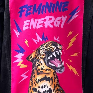 Può includere: Giacca di jeans nera con una toppa rosa vibrante con la scritta "FEMININE ENERGY" in blu e rosa. Una grafica di leopardo ruggente è circondata da fulmini gialli e blu.