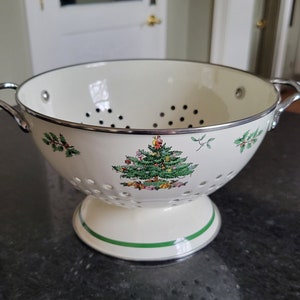 May include: White enamel colander with a green and red Christmas tree design. The colander has a stand and two handles.