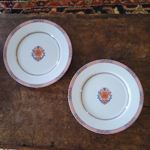 May include: Two white ceramic plates with a blue and orange floral design. The plates have a gold trim and are on a wooden table.