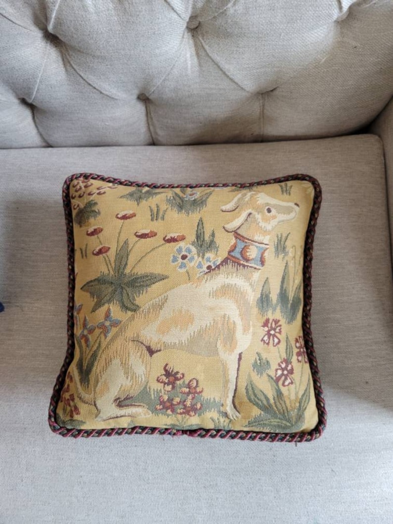 Puede incluir: Una almohada cuadrada con un ribete trenzado marr&oacute;n y rojo. La almohada presenta un dise&ntilde;o de tapicer&iacute;a de un perro blanco con un collar azul de pie en un campo de flores amarillas.