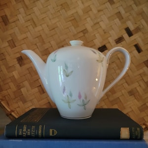 May include: A white porcelain teapot with a floral design and a gold rim. The teapot is sitting on top of two books, one blue and one black. The books have text in Japanese and German.