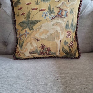 Puede incluir: Una almohada decorativa con un dise&ntilde;o de tapiz que presenta un perro blanco con un collar azul y dorado en un campo de flores. La almohada tiene un ribete trenzado marr&oacute;n y rojo.