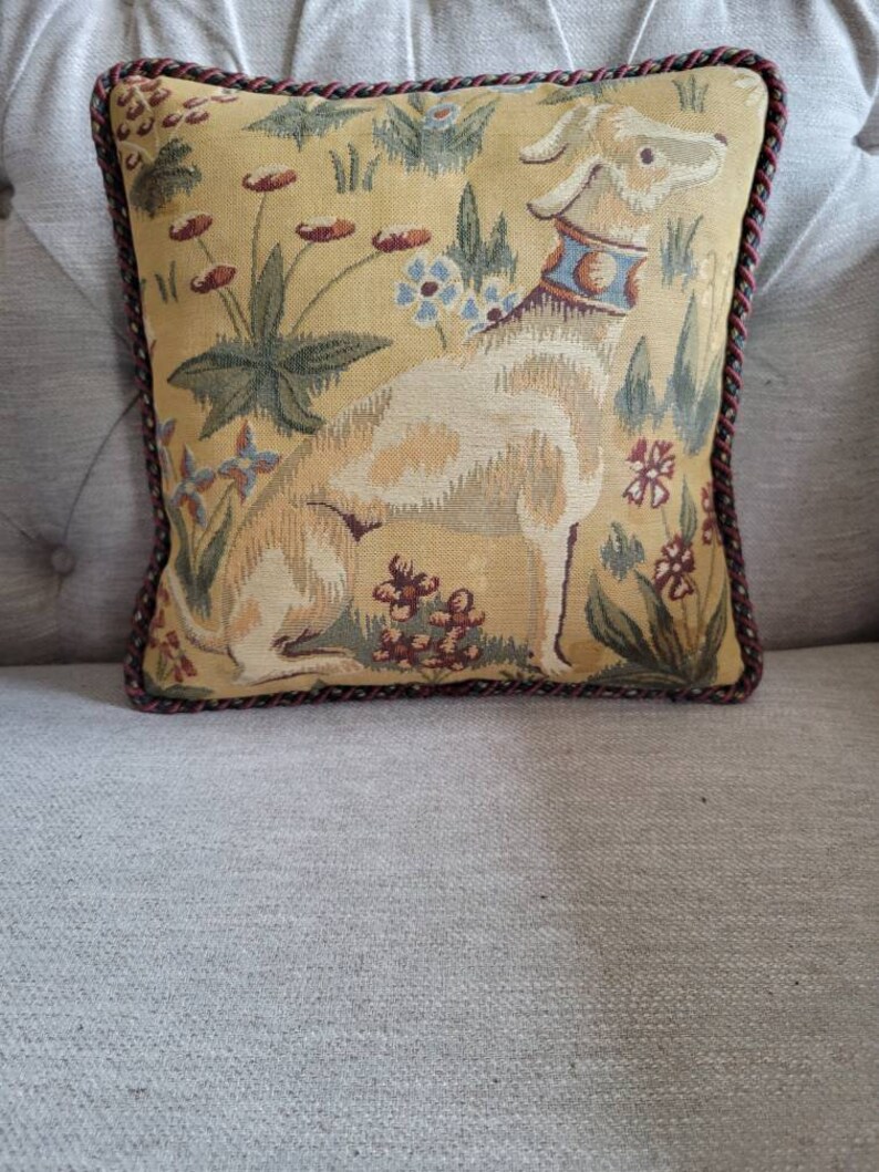 Puede incluir: Una almohada decorativa con un dise&ntilde;o de tapiz que presenta un perro blanco con un collar azul y dorado en un campo de flores. La almohada tiene un ribete trenzado marr&oacute;n y rojo.