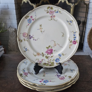 May include: A stack of antique porcelain plates with floral designs. The plates have a cream-colored base, adorned with pink, blue, and purple flowers. Each plate has a gold trim around the edge. The plates are displayed on a dark wooden surface.