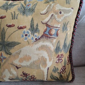 Puede incluir: Una almohada decorativa con un dise&ntilde;o de tapiz que presenta un perro blanco con un collar azul y dorado, rodeado de patrones florales verdes y marrones sobre un fondo beige. La almohada tiene un ribete trenzado rojo y marr&oacute;n.