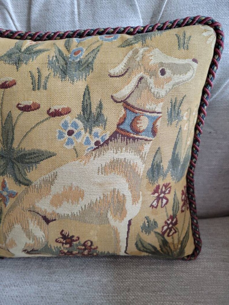 Puede incluir: Una almohada decorativa con un dise&ntilde;o de tapiz que presenta un perro blanco con un collar azul y dorado, rodeado de patrones florales verdes y marrones sobre un fondo beige. La almohada tiene un ribete trenzado rojo y marr&oacute;n.