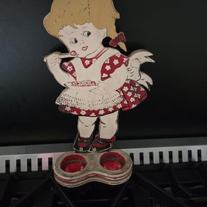 May include: A vintage wooden figurine of a girl with blonde hair, wearing a red and white dress with floral accents. The figurine is standing on a double-holed, red-rimmed holder. The girl is holding a spoon, and the figurine has a hole at the top for hanging.