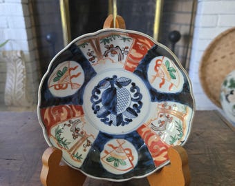 May include: A decorative, scalloped-edge plate with a detailed, colorful design. The plate features a central blue and white pattern, surrounded by alternating sections of red, white, and blue designs. It is displayed on a wooden stand.