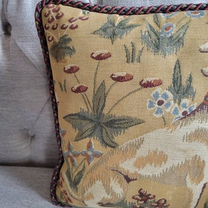 Puede incluir: Una almohada decorativa con un dise&ntilde;o de tapiz que presenta un perro blanco, hojas verdes y flores rojas y azules sobre un fondo amarillo. La almohada tiene un ribete trenzado rojo y verde.