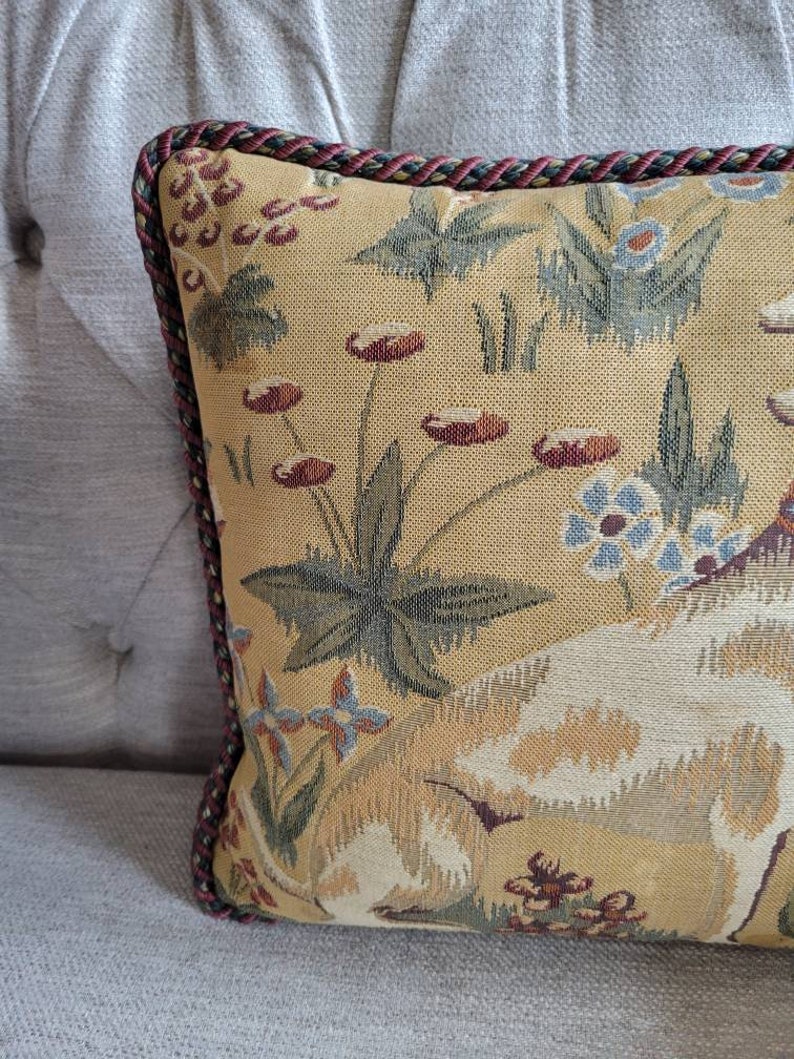 Puede incluir: Una almohada decorativa con un dise&ntilde;o de tapiz que presenta un perro blanco, hojas verdes y flores rojas y azules sobre un fondo amarillo. La almohada tiene un ribete trenzado rojo y verde.