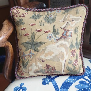 Puede incluir: Una almohada beige con un dise&ntilde;o de tapicer&iacute;a que presenta un perro blanco y patrones florales. La almohada tiene un ribete trenzado rojo y marr&oacute;n.