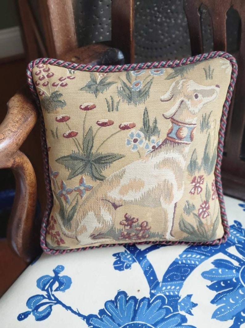 Puede incluir: Una almohada beige con un dise&ntilde;o de tapicer&iacute;a que presenta un perro blanco y patrones florales. La almohada tiene un ribete trenzado rojo y marr&oacute;n.