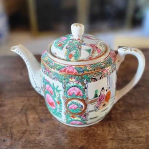 May include: A white porcelain teapot with a lid, decorated with a colorful floral and geometric pattern. The teapot has a gold trim and features a scene of people in traditional Chinese clothing.