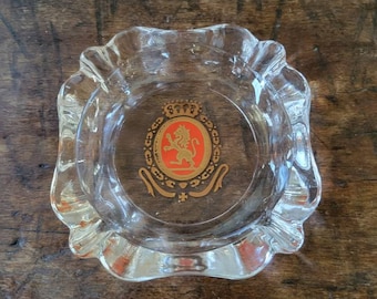 May include: A clear glass ashtray with a gold and red crest featuring a lion. The crest has a crown and a banner with the words "C.C.C.S."