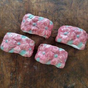 May include: Four pink ceramic napkin rings with a floral design. The rings are decorated with pink flowers and green leaves.