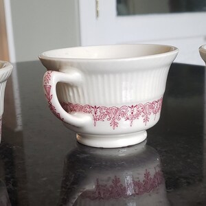 May include: Three white teacups with pink floral designs around the rim. The cups have a fluted design and a handle.