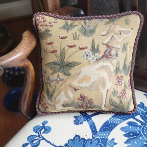 Puede incluir: Una almohada cuadrada con un dise&ntilde;o de tapiz que presenta un perro blanco en un campo de flores. La almohada tiene un ribete trenzado rojo y marr&oacute;n.