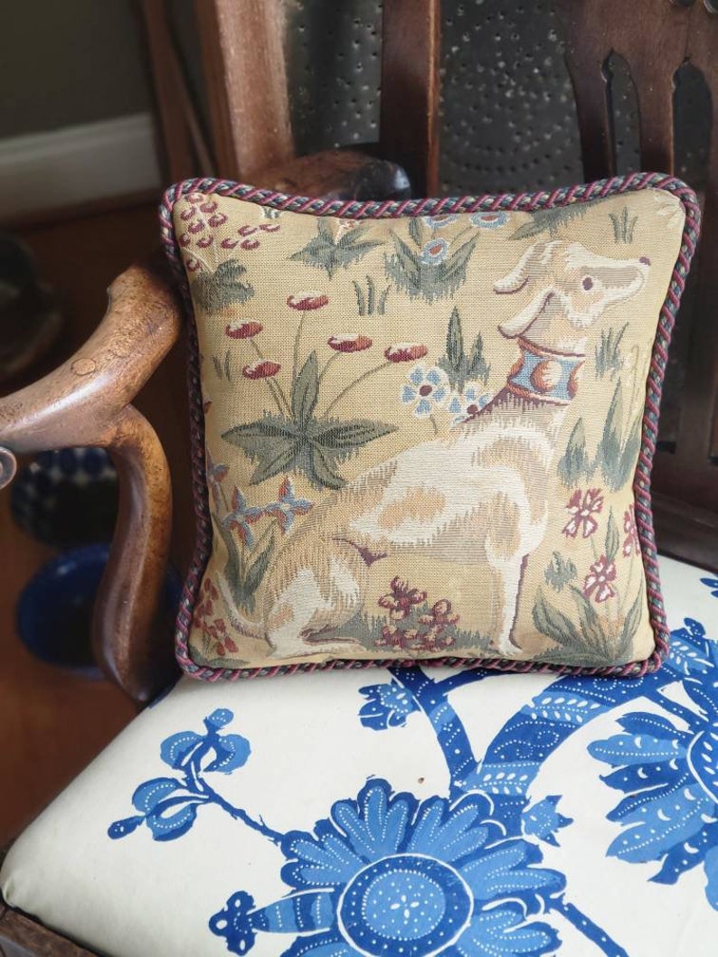 Puede incluir: Una almohada cuadrada con un dise&ntilde;o de tapiz que presenta un perro blanco en un campo de flores. La almohada tiene un ribete trenzado rojo y marr&oacute;n.