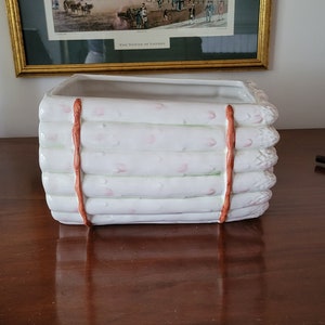 May include: White ceramic planter shaped like a bundle of asparagus with brown ties. The planter has a rectangular shape and is decorated with pink flowers.