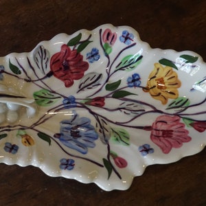 May include: A white ceramic leaf-shaped serving dish with a floral design. The dish features pink, yellow, blue, and green flowers with a white background.
