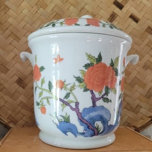 May include: White porcelain jar with a lid, featuring a floral design with orange flowers and blue and green leaves. The jar has two handles on the sides.