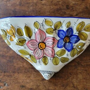 May include: A white ceramic wall pocket with a blue rim and a floral design. The flowers are pink, blue, and yellow with green leaves.