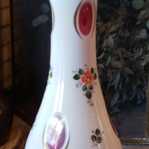 May include: White glass decanter with a pink and gold design. The decanter has a round, clear glass stopper with a gold band. The decanter is decorated with painted flowers and red and gold accents.