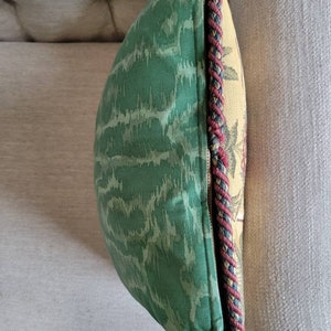 Puede incluir: Una almohada de tiro verde y crema con un patr&oacute;n tejido y un ribete trenzado rojo, negro y crema.