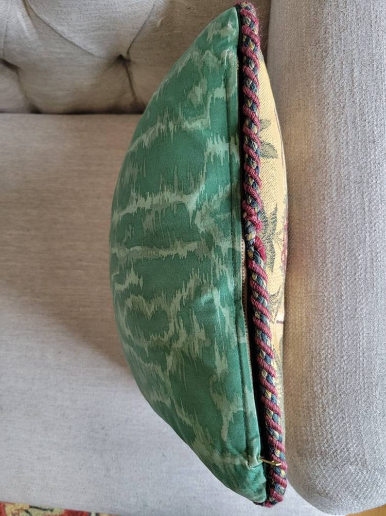 Puede incluir: Una almohada de tiro verde y crema con un patr&oacute;n tejido y un ribete trenzado rojo, negro y crema.