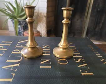 May include: Two gold metal candle holders with a simple design. The candle holders are sitting on a green book with the text "Ralph Lauren in his own fashion" on the cover.