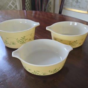 May include: Three white ceramic baking dishes with a yellow glaze and green floral pattern. The dishes are on a brown wooden table.