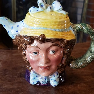 May include: A ceramic teapot shaped like a woman's head wearing a yellow hat with blue polka dots. The teapot has a green handle and a blue and white polka dot spout.