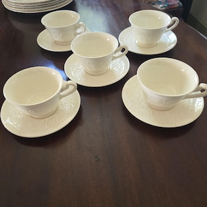 May include: A set of six white teacups and saucers with a floral embossed design. Additional matching plates are stacked in the background. The teacups have a curved handle and sit on round saucers.