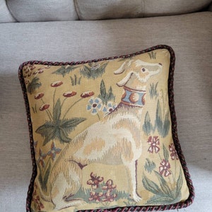 Puede incluir: Una almohada cuadrada con un dise&ntilde;o de tapiz que presenta un perro blanco sentado en un campo de flores. La almohada tiene un ribete trenzado marr&oacute;n y dorado.