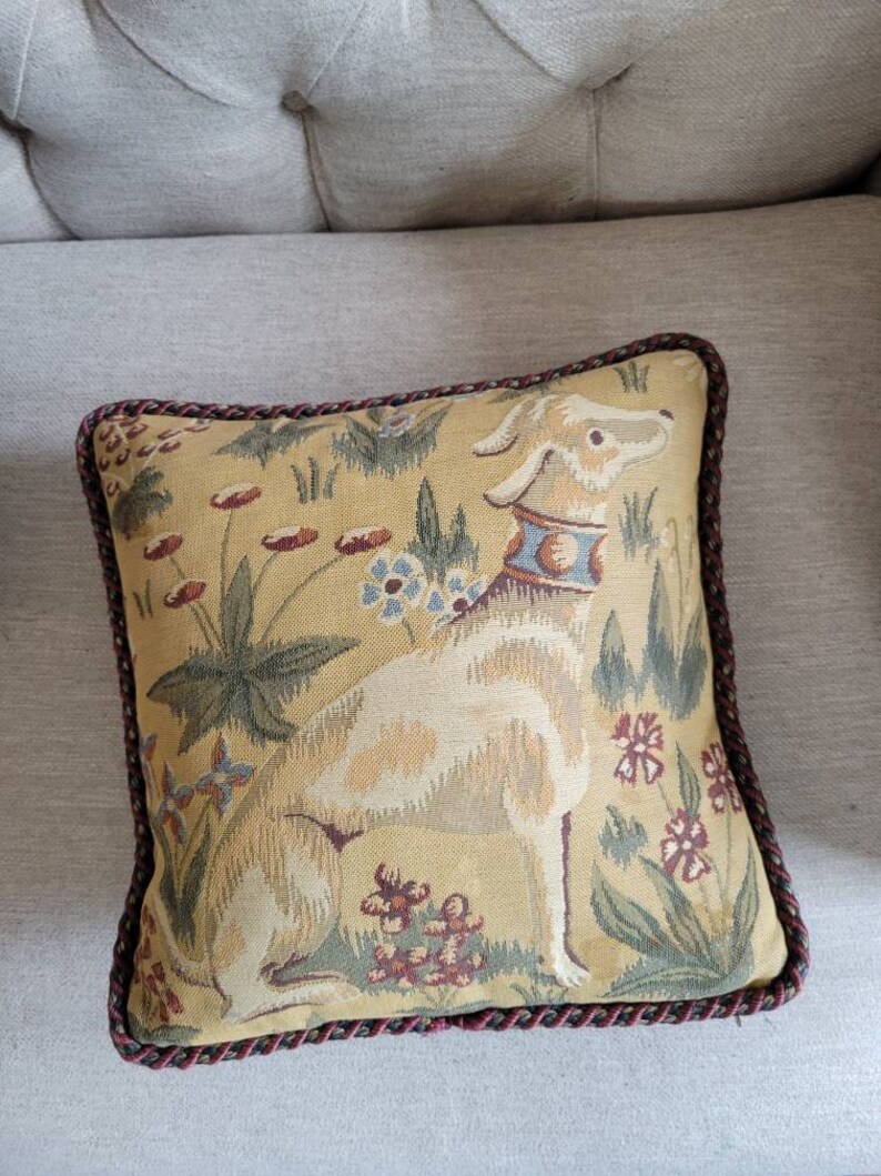 Puede incluir: Una almohada cuadrada con un dise&ntilde;o de tapiz que presenta un perro blanco sentado en un campo de flores. La almohada tiene un ribete trenzado marr&oacute;n y dorado.