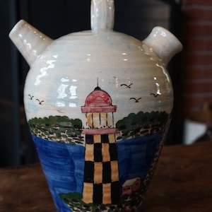 May include: A white ceramic pitcher with a lighthouse painted on the front. The lighthouse is black and yellow with a red roof. The pitcher has two spouts and a handle.