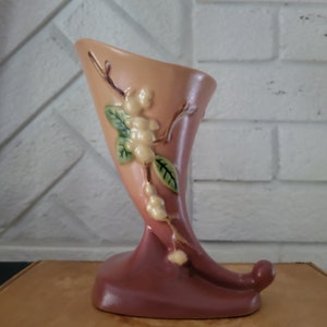 May include: A pink and brown ceramic vase shaped like a horn with a floral design. The vase has a white and green floral design with a branch and leaves.