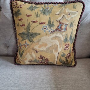 Puede incluir: Una almohada decorativa con un dise&ntilde;o de tapiz que presenta un perro beige con un collar azul y un patr&oacute;n floral sobre un fondo amarillo. La almohada tiene un ribete trenzado marr&oacute;n y rojo.